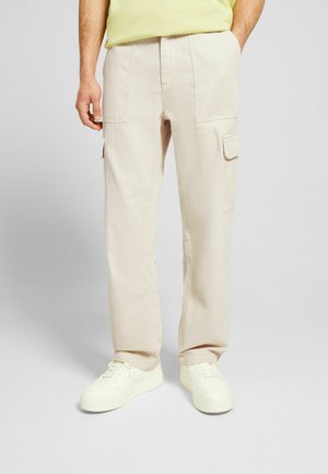 Homme portant un pantalon cargo beige avec des poches et des baskets blanches, debout devant un fond uni.
