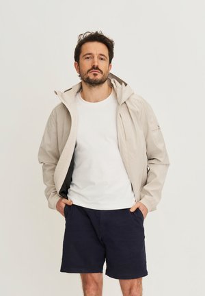 Man met baard, gekleed in een beige jas over een wit T-shirt en marineblauwe korte broek, staand met handen in de zakken tegen een effen achtergrond.