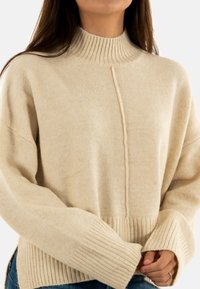 Pull en tricot de couleur crème avec un col haut à côtes, des épaules tombantes et des poignets à côtes. Présente un détail de couture central. Texture douce.