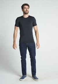 Getailleerde zwarte t-shirt met korte mouwen, gecombineerd met donkerblauwe jeans. Model staat rechtop voor een effen achtergrond en draagt casual blauwe schoenen.