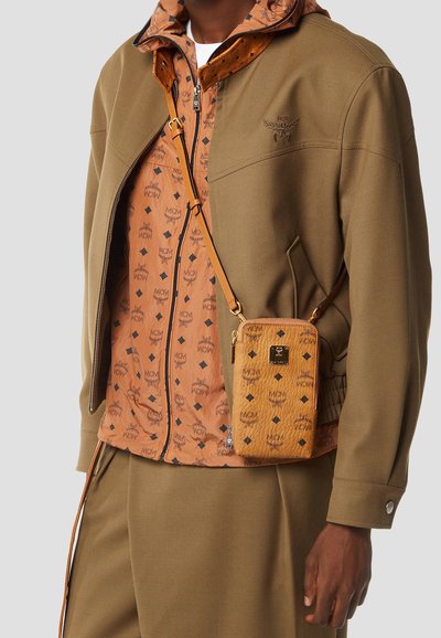 Homme portant une veste beige et un pantalon assorti avec un sweat à capuche zippé à motifs orange et un petit sac bandoulière beige.
