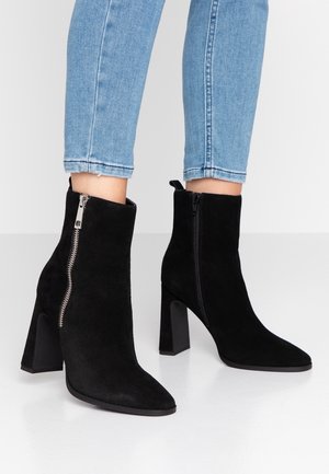 Schwarze Wildleder-Stiefeletten mit Blockabsätzen und Reißverschlüssen auf hell gebleichten blauen Jeans vor einfarbig weißem Hintergrund.