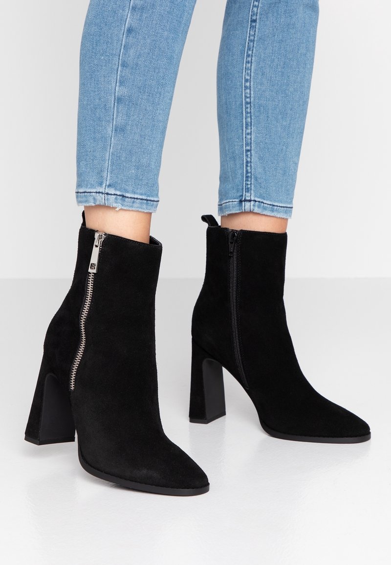 Bottines en daim noir à talons blocs avec fermetures éclair portées sur un jean en denim bleu délavé clair, sur fond blanc uni.