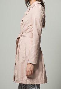 Ljusrosa trenchcoat med bälte i midjan, skräddarsydd krage och långa ärmar. Har två sidofickor och ett slätt, något glänsande tyg.