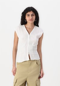 Haut blanc à manches courtes, avec un décolleté en V et une fermeture à boutons, présentant un tissu texturé et une coupe ajustée avec des détails de couture sur le devant.