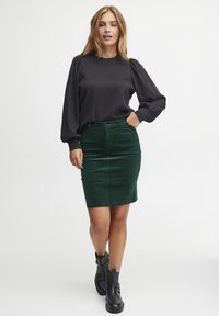 Crna bluzica s puff rukavima u paru s tamnozelenom kordura suknjom olovkom. Model nosi crne gležnjače, ističući prilagodljiv siluetu i teksturu.
