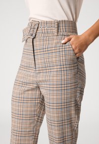 Beige geruite broek met een hoge taille, geaccentueerde riem en zijzakken. De stof heeft een ruitpatroon in bruin en marineblauwe tinten.