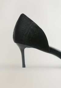 Czarne aksamitne szpilki z wąskim noskiem i smukłym, średniej wysokości obcasem w stylu stiletto na jasnym, neutralnym tle.