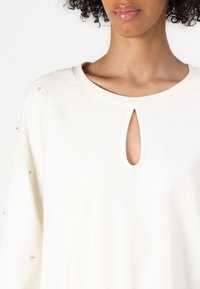 Vrouw draagt een wit shirt met lange mouwen, een keyhole-halslijn en decoratieve knopen langs de mouw, tegen een effen achtergrond.
