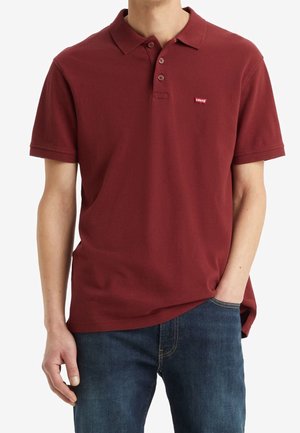 Mężczyzna w bordowej koszulce polo z krótkim rękawem z logo Levi's na piersi i niebieskich dżinsach, z ręką w kieszeni, stojący na białym tle.