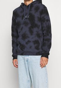 Felpa con cappuccio tie-dye blu navy, dotata di tasca a marsupio, cappuccio con cordoncino e polsini neri. Presenta un piccolo logo bianco ricamato sul petto.