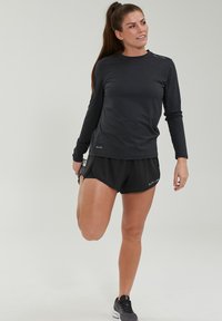 Haut de sport noir à manches longues associé à un short noir ; possède un col rond, une texture douce et un logo minimaliste.