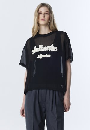 Młoda kobieta ubrana w przezroczysty czarny top z napisem "Authentic Letters" na czarnym topie na ramiączkach, w połączeniu z luźnymi ciemnoszarymi spodniami.