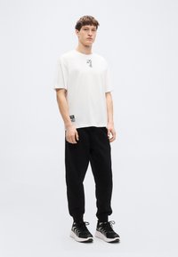 Λευκό βαμβακερό T-shirt με μαύρο αφηρημένο γραφικό στο στήθος, συνδυασμένο με μαύρο παντελόνι jogger και μαύρα αθλητικά παπούτσια με κυματιστό σχέδιο.