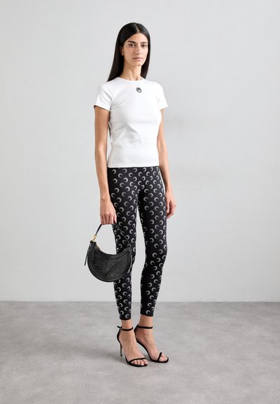 Hvid tætsiddende t-shirt med et rundt logo, parret med sorte leggings med et gentagende halvmåne-mønster og en sort halvmåne-formet håndtaske.