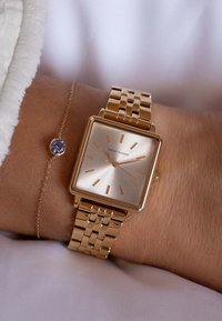 Gouden metalen horloge met een rechthoekige wijzerplaat, lichte gouden wijzerplaat en dunne uurmarkeringen. Heeft een gelinkte band en een delicate armband met een paarse edelsteen.