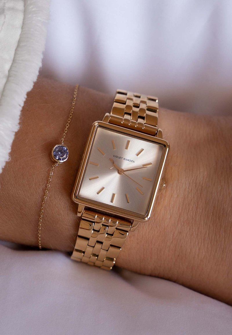 Gouden metalen horloge met een rechthoekige wijzerplaat, lichte gouden wijzerplaat en dunne uurmarkeringen. Heeft een gelinkte band en een delicate armband met een paarse edelsteen.