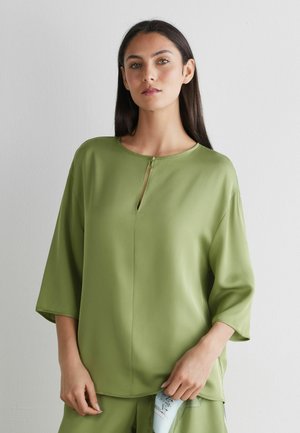 Kvinde med langt mørkt hår iført en lysegrøn, løs bluse med en lille knapåbning ved halsudskæringen, poserende mod en ensfarvet hvid væg.