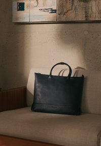 Geantă tote din piele neagră, cu două mânere, design elegant, suprafață texturată, expusă pe o canapeabej, în fața unui perete de piatră.