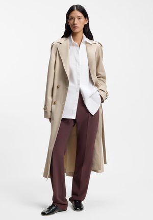 Ženska, oblečena v bež trench coat čez belo srajco s gumbi in bordo hlače, stoji z eno roko v žepu na belem ozadju.