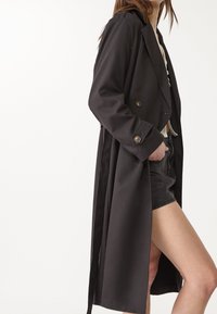Langer schwarzer Trenchcoat mit doppelreihigem Design, großen Knöpfen und glattem Stoff. Verfügt über einen gebundenen Gürtel und seitliche Schlitze für Bewegungsfreiheit.