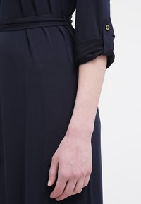 Marinblå klänning med uppkavlade ärmar, som har ett bälte i midjan och detaljer med guldknappar. Den släta texturen och den skräddarsydda passformen framhäver silhuetten.