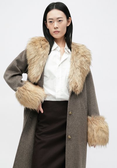 Καφέ μάλλινο παλτό με γιακά και μανίκια από συνθετική γούνα. Διαθέτει χρυσά κουμπιά και ευθεία γραμμή. Φοριέται πάνω από ένα λευκό πουκάμισο και μια σκούρα φούστα.