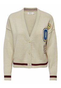 Beżowy sweter robiony na drutach z głębokim dekoltem w kształcie litery V, zapinany na guziki, z bordowymi paskami. Posiada haftowane naszywki, w tym uśmiechniętą buźkę.