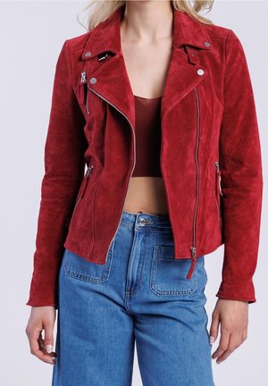 Mujer con una chaqueta biker de ante roja con cremalleras plateadas sobre un top corto y vaqueros azules de cintura alta sobre un fondo liso.