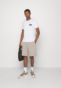 T-shirt bianca con un piccolo etichetta nera, pantaloni corti beige e scarpe da ginnastica nere. I pantaloni corti hanno un logo sottile, mentre la borsa è nera e testurizzata.