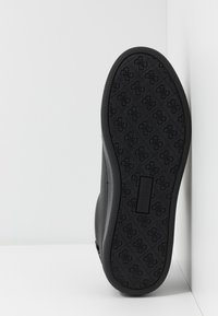 Semelle de chaussure en caoutchouc noir présentant une texture texturée et un logo embossé au centre ; inclinée sur un fond gris clair.