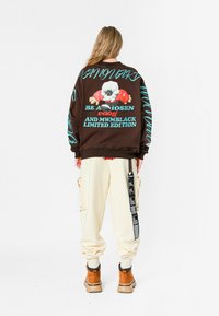 Chaqueta bomber marrón con gráficos coloridos, corte oversize, puños acanalados, combinada con pantalones cargo crema, botas naranjas y un detalle de correa negra.
