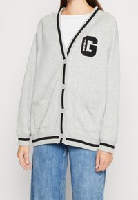 Cardigan gris claro con ribete negro, cinco botones, dos bolsillos frontales y un parche con la letra "G" que presenta el año "1969" en la parte izquierda del pecho.