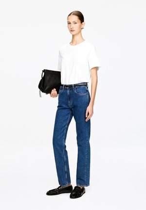 Mujer con camiseta blanca de manga corta, vaqueros azules de tiro alto, cinturón negro, mocasines negros, sosteniendo un bolso de mano de cuero negro.