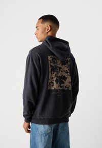 Sort hættejacke med snøre, der har et stort brunt tie-dye print og tekstdesign på ryggen. Blødt materiale, ribbede manchetter og kant.