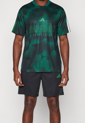 Mężczyzna w zielono-czarnej moro koszulce sportowej Adidas i czarnych szortach Adidas z zielonym logo, stojący na jednolitym tle.