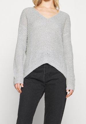 Mujer que lleva un suéter de punto gris claro con cuello en V y mangas largas y pantalones negros de talle alto sobre un fondo liso.