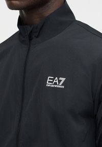 Μαύρο τζάκετ με φερμουάρ που φοριέται από άτομο με σκουρόχρωμο δέρμα, με λευκό λογότυπο EA7 Emporio Armani στο αριστερό μέρος του στήθους.