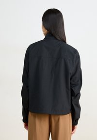 Svart vindjacka med hög krage, elastiska ärmmuddar och en avslappnad passform. Har en liten logotyp på övre ryggen.