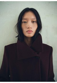 Manteau en laine bordeaux avec un large col cranté et une fermeture à boutons, affichant une silhouette structurée et une texture lisse.
