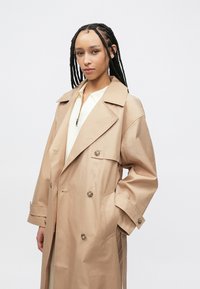 Μπεζ διπλής σειράς καμπαρντίνα με μεγάλα πέτα, κλεισίματα με κουμπιά και πλαϊνές τσέπες. Κατασκευασμένη από λείο, ελαφρύ ύφασμα.