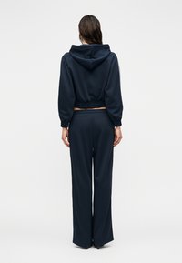 Μπλε ναυτικού χρώματος φούτερ με κουκούλα, ελαστική μέση και μανσέτες, συνδυασμένο με παντελόνια φαρδιάς γραμμής. Λείο ύφασμα με ελάχιστες λεπτομέρειες, που τονίζει ένα casual σχέδιο.