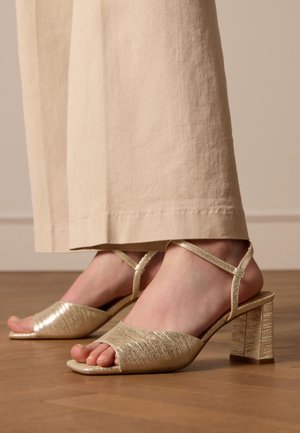 Gold, strappy heeled sandals with a peep toe. Textured upper, chunky heel, and slender ankle straps. Woodgrain pattern on the heel.