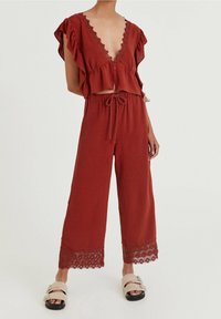Roodbruine twee-delige outfit met een cropped, gerimpeld blouse met kanten afwerking en een broek met wijdere pijpen, voorzien van kantaccenten aan de zoom.