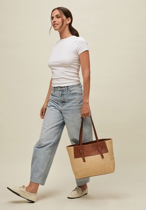 Donna che cammina con una grande borsa intrecciata con dettagli in pelle marrone, indossa una t-shirt bianca, jeans azzurri e mocassini crema.