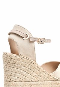 Sandale compensée espadrille beige avec une base en jute tissée texturée, un dessus en daim et une bride de cheville réglable avec une quincaillerie en métal.