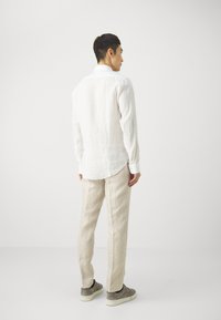 Camisa de lino blanca de manga larga, con puños abotonados y un dobladillo curvado, combinada con pantalones de lino beige claro y zapatillas grises.