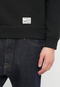 Κοντινό πλάνο ενός ατόμου που φοράει μαύρο φούτερ με λευκή ετικέτα πατσά και σκούρο μπλε τζιν με ορατές κίτρινες ραφές.