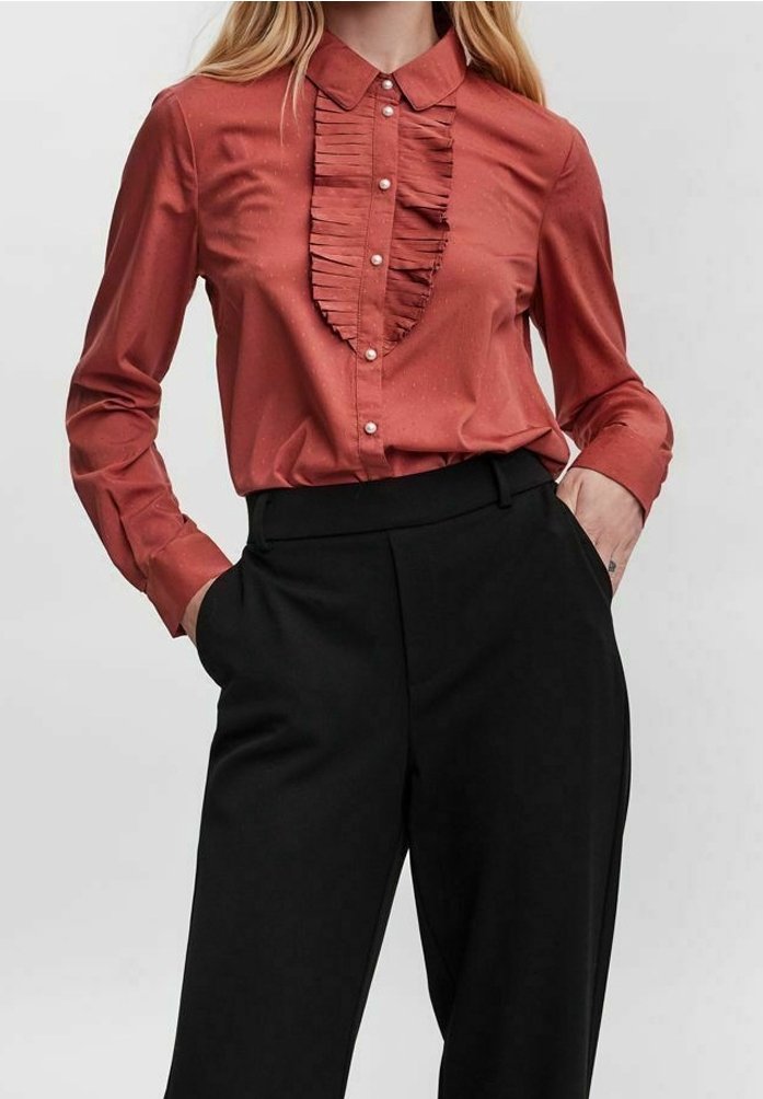 Blouse de couleur rouille avec devant à volants et boutons en perle, associée à un pantalon noir taille haute, mains dans les poches.