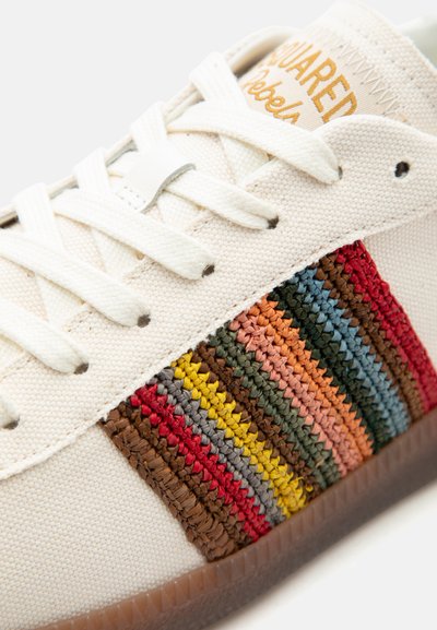 Κοντινό πλάνο λευκού αθλητικού παπουτσιού με πολύχρωμες υφαντές ρίγες, λευκά κορδόνια και το κέντημα "Dsquared Rebels" με πορτοκαλί γράμματα στη γλώσσα του παπουτσιού.